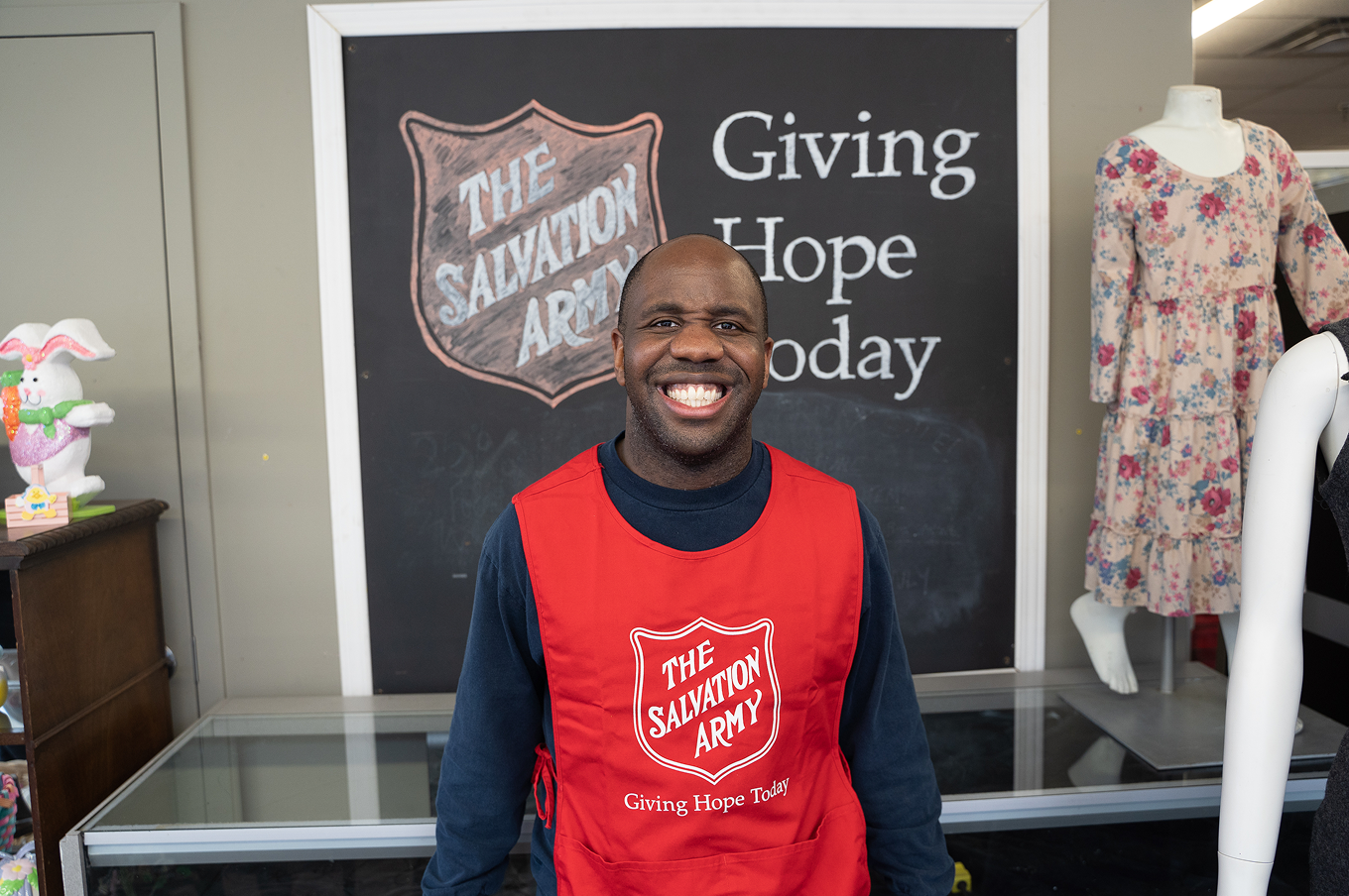 Smiling Salvation Army staff member wearing a red vest inside a thrift store, creating a welcoming atmosphere.